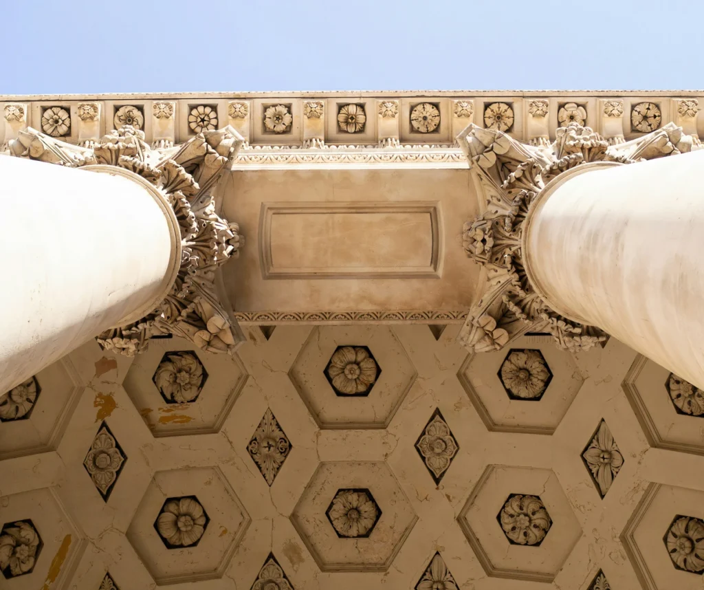 Ornate architectural columns and ceiling detail with intricate floral patterns against a clear blue sky.