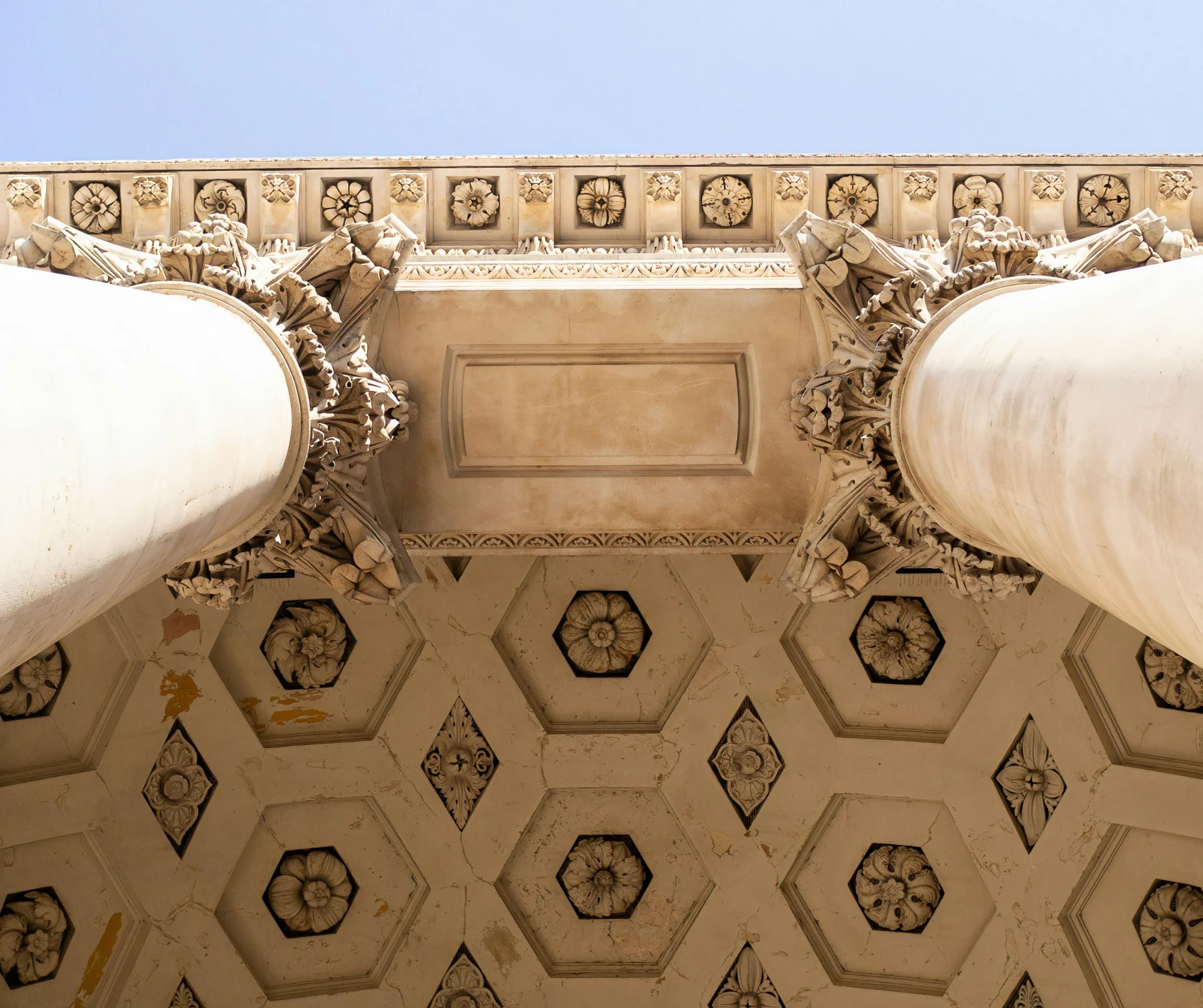 Ornate architectural columns and ceiling detail with intricate floral patterns against a clear blue sky.